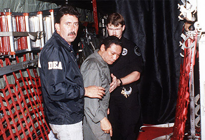 Manuel Noriega is escorted by U.S. Drug Enforcement Agency personnel aboard an aircraft at Howard Air Force Base, Panama, after turning himself over on 3 January 1990. 