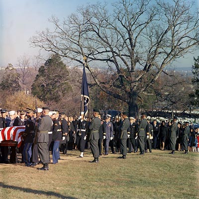Special Forces line the path between the road and the gravesite at Arlington as JFK's casket passes by. Jacqueline and Robert Kennedy follow behind, towards the right side of the photo.