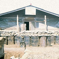 Team 9’s house at Edap Enang. In front of the quarters are barrels filled with earth and topped with sandbags for protection against VC attacks.