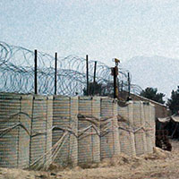 Photo showing a portion of the HESCO perimeter wall around the CJSOTF Headquarters in April 2002. Note the addition of lighting for night operations.  Surveillance cameras were installed to allow guards to remotely view sectors without exposing themselves to direct fire weapons.