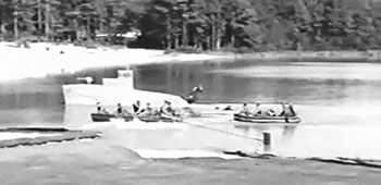 Special Forces wearing SCUBA gear jump into McKellar’s Pond from a plywood silhouette patrol boat while an SF team paddles alongside in RB-7 rubber boats.