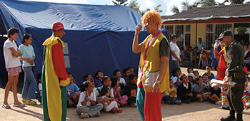 Children listen to a presentation given by the Colombian National Police while their parents wait in line for other activities. The police officers dressed as clowns are used to make the counter-drug message entertaining.