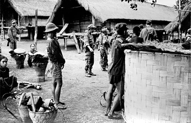 Patrol in a Montagnard village. In the center of the photo is a VC prisoner (dressed in shorts and a “boonie cap” with his hands tied in front). The striker on the right is leaning on the village communal latrine.
