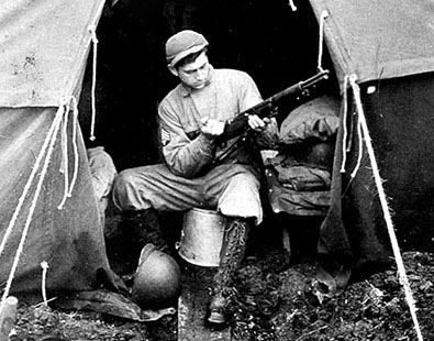 A Forceman cleans his M-1 rifle in a Bell tent on Amchitka. His knee-high shoepacs and the improvised boardwalk helped to traverse the soft muskeg of the island.