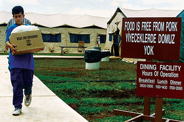 A Kurdish refugee boy carries food for his family to his temporary home at Andersen AFB, Guam from the dining facility. The sign on the right assures the Kurds that the food is “Free from Pork,” meeting Muslim religious dietary customs. 