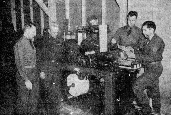 LTC Frank McCulloch presses the “Start” button as CPT Otto Bennett looks on. PVT Rus, 1LT Peter A. DeFranco, and SGT Gilbert examine the first copies of an offset press-printed <i>Psyn-post</i> in Building 193, Fort Riley.