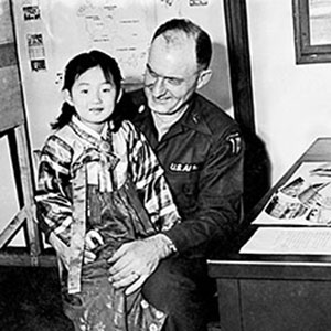A Korean girl sits on the lap of Lieutenant Colonel Jack T. Shannon, who served first as the Psywar officer, then the Chief of the Civil Information Section with KCAC from October 1954 to April 1955 (note the KCAC SSI).