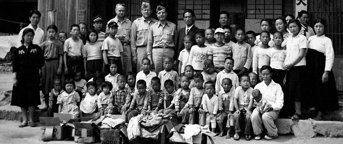American officers of the 1903rd Engineer Aviation Battalion pose with children and staff of the Tae Jee Orphanage in 1952 with boxes of donated clothing.