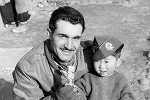 Obviously missing his twin sons back home SFC James C. ‘Joe’ Pagnella has perched his overseas cap on a little Korean boy more interested in his  pack of Necco Jells.