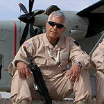 CW5 Arooji sitting in front of a plane alongside two other soldiers.